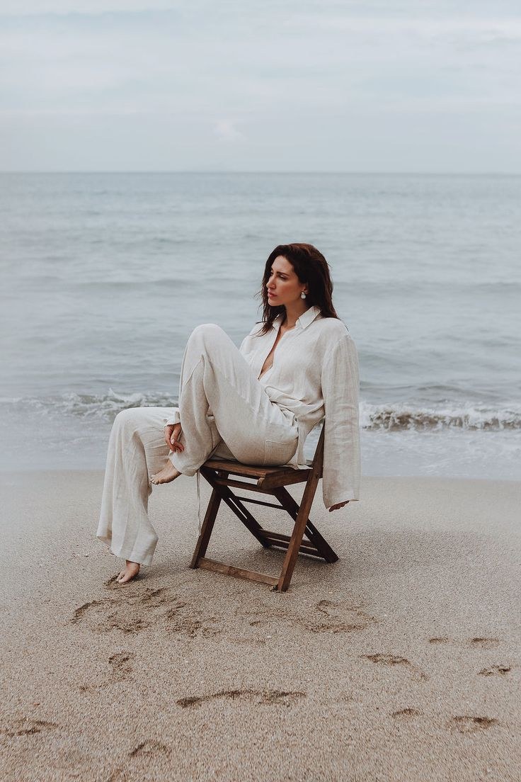 Woman in a cream linen shirt and trousers seated on a wooden chair on the beach